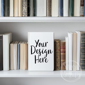 Peut inclure: Une maquette de livre blanc sur une étagère blanche avec une variété de livres de chaque côté. Le livre a le texte "Your Design Here" écrit sur la couverture.