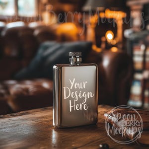 May include: A silver hip flask with a screw-on top. The flask has the text "Your Design Here" engraved on the front. The flask is sitting on a wooden table with a brown leather journal.