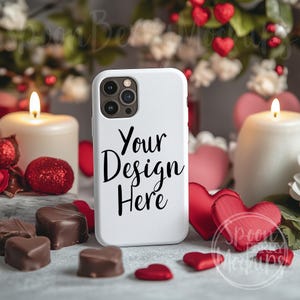 May include: White iPhone case mockup with the text "Your Design Here" on a white surface with red hearts, candles, and chocolate hearts.