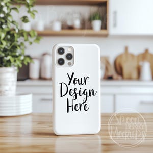 Puede incluir: Una funda de teléfono blanca con el texto "Your Design Here" en escritura negra. La funda tiene un módulo de cámara con múltiples lentes. Está sobre una superficie de madera, con un fondo de cocina borroso.