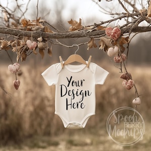May include: White baby onesie mockup hanging from a branch with brown leaves and red heart-shaped decorations. The onesie has the text "Your Design Here" printed on the front.