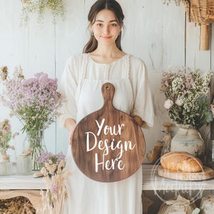 Könnte beinhalten: Eine Frau in einem weißen Kleid hält ein rundes, hölzernes Schneidebrett mit dem Text "Your Design Here" in weißen Buchstaben. Das Brett hat einen Griff und ist perfekt, um ein Design oder Logo zu präsentieren.
