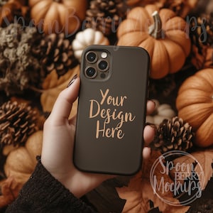 May include: A black phone case with a white text overlay that says "Your Design Here". The phone case is being held by a hand in front of a background of fall-themed decorations, including pumpkins and pine cones.