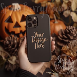 Puede incluir: Una funda de teléfono negra con una inscripción dorada que dice "Your Design Here". La funda está siendo sostenida por una mano frente a un fondo de decoraciones de otoño, incluyendo calabazas y piñas.