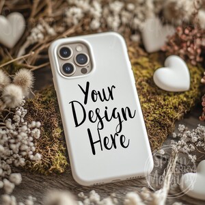 May include: White phone case mockup featuring a camera lens cutout and the text "Your Design Here" in black script font. The case is resting on a bed of moss and white flowers.