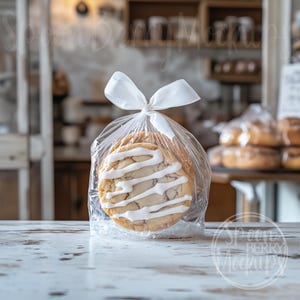May include: Two sugar cookies with white icing are packaged in a clear plastic bag tied with a white ribbon. The cookies are on a white wooden table.
