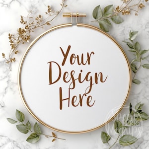 Puede incluir: Un aro de bordado dorado con un lienzo blanco y el texto "Your Design Here" en letras marrones. El aro está rodeado de follaje verde y dorado sobre un fondo de mármol blanco.