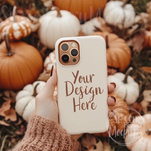 May include: A white iPhone case with a gold camera lens and the text "Your Design Here" in brown lettering. The case is being held by a hand in front of a background of pumpkins and gourds.