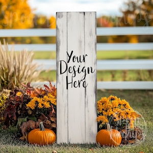 Könnte beinhalten: Ein weißes Holzschild mit einer abgenutzten Oberfläche, das vor einem weißen Lattenzaun steht. Das Schild trägt den Text "Your Design Here" in Schwarz. Das Schild ist von Herbstlaub umgeben, darunter Kürbisse und gelbe Blumen.