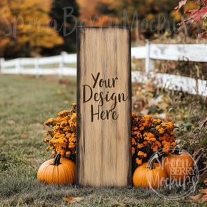 May include: A rustic wooden sign with the text "Your Design Here" in brown lettering. The sign is standing in front of a white picket fence with pumpkins and fall foliage in the foreground.