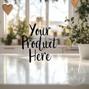May include: A bright, airy kitchen scene with a marble countertop. The text "Your Product Here" is displayed in black script. A potted plant and gold glitter heart ornaments add decorative touches. The background features a window and greenery.