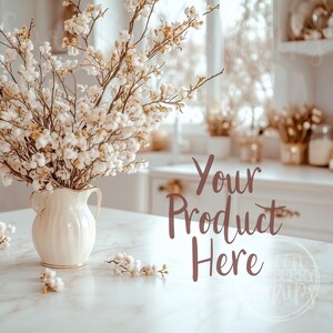 Puede incluir: Una jarra de cerámica blanca llena de flores blancas secas se encuentra sobre una encimera de mármol blanco. El texto "Your Product Here" está escrito en cursiva marrón sobre la encimera.
