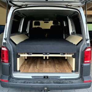 Pode incluir: Interior de carrinha de campismo personalizado com estrutura de cama e colchão. A estrutura da cama é feita de madeira clara e o colchão é preto. A carrinha tem piso com aspecto de madeira. As portas traseiras estão abertas, revelando o interior.