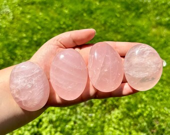 Può includere: Quattro pietre di quarzo rosa lisce, di forma ovale e di diverse dimensioni, con una tonalità rosa e striature bianche, tenute in una mano su uno sfondo verde.