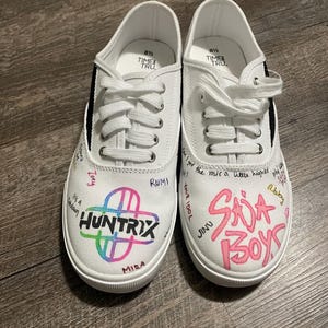Puede incluir: Un par de zapatillas de lona blancas con detalles negros y cordones blancos. La zapatilla izquierda presenta la palabra "HUNTRIX" en un diseño colorido, y la derecha dice "SADA BOYS" en rosa. Ambas zapatillas tienen texto escrito a mano.