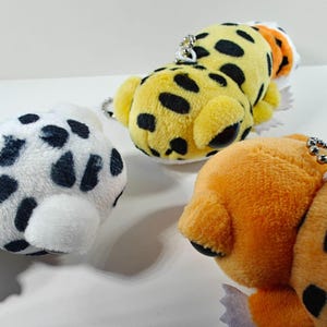 May include: Close-up of three plush animal keychains. One is white with black spots, another is yellow with black spots, and the third is orange with black spots. Each has a silver chain for attachment.