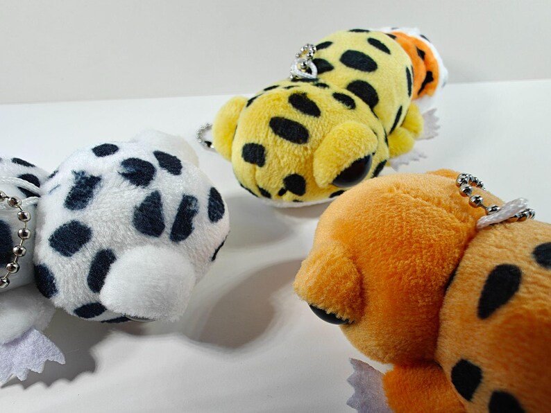 May include: Close-up of three plush animal keychains. One is white with black spots, another is yellow with black spots, and the third is orange with black spots. Each has a silver chain for attachment.