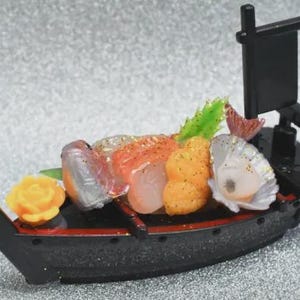 Op de afbeelding: Een miniatuur sushi boot, zwart met rode accenten, beladen met kleurrijke sushi. De sushi omvat oranje, witte en roze vis, een gele bloem en groene garnering. Een kleine zwarte vlag staat achterop.