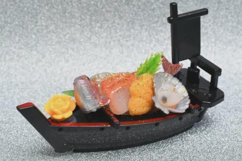 Op de afbeelding: Een miniatuur sushi boot, zwart met rode accenten, beladen met kleurrijke sushi. De sushi omvat oranje, witte en roze vis, een gele bloem en groene garnering. Een kleine zwarte vlag staat achterop.