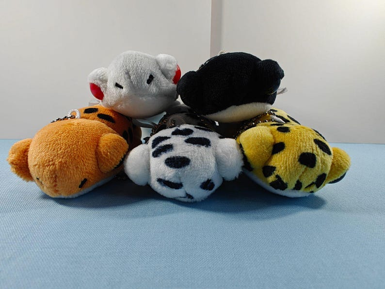 May include: A collection of small, plush animal head keychains. The keychains include a white bear with red ears, an orange tiger, a black bear, a white dalmatian, and a yellow leopard with black spots.