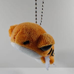 May include: A plush orange and white tiger keychain with black stripes and large black eyes. The small stuffed animal is attached to a silver ball chain. The tiger has a white mouth and a small white tongue.