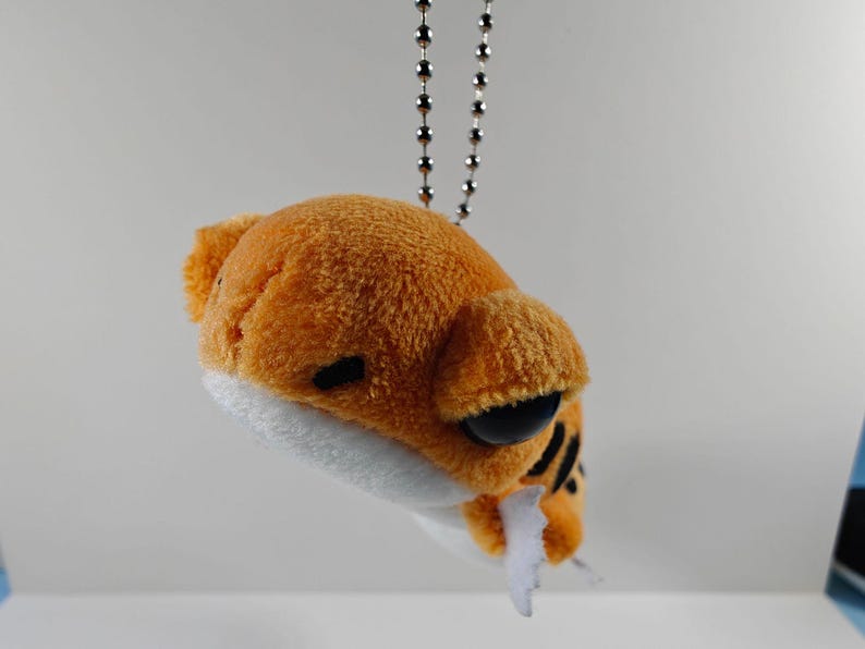 May include: A plush orange and white tiger keychain with black stripes and large black eyes. The small stuffed animal is attached to a silver ball chain. The tiger has a white mouth and a small white tongue.