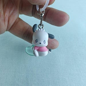 Puede incluir: Un peque&ntilde;o llavero de perro de dibujos animados blanco y rosa con orejas negras y una camisa rosa. El llavero est&aacute; sujeto a un cierre plateado y a un dije transparente en forma de coraz&oacute;n. Una presilla blanca est&aacute; sujeta en la parte superior.