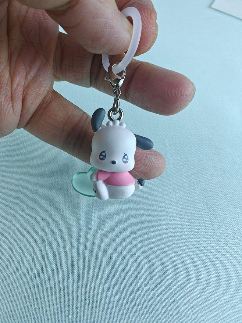 Puede incluir: Un peque&ntilde;o llavero de perro de dibujos animados blanco y rosa con orejas negras y una camisa rosa. El llavero est&aacute; sujeto a un cierre plateado y a un dije transparente en forma de coraz&oacute;n. Una presilla blanca est&aacute; sujeta en la parte superior.