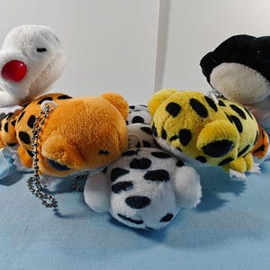 May include: A collection of plush animal keychains in various designs. Includes a white bird with a red beak, orange and yellow spotted animals, a black and white frog, and a dalmatian dog. Each keychain has a silver chain.