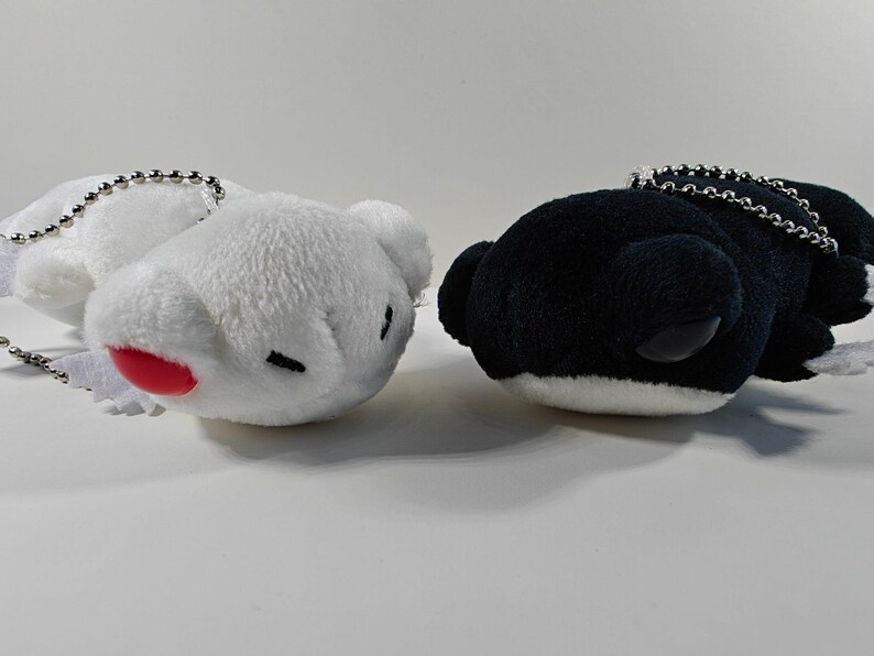 May include: Two plush whale-shaped keychains. One is white with a red mouth and black eyes, the other is black with white accents. Both have silver chain attachments.
