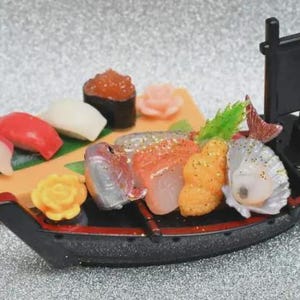 Op de afbeelding: Een miniatuur sushi boot met diverse sushi stukken, waaronder nigiri en sashimi. De boot is zwart met rode accenten, en de sushi heeft levendige kleuren zoals rood, oranje en wit. Achteraan staat een kleine zwarte vlag.