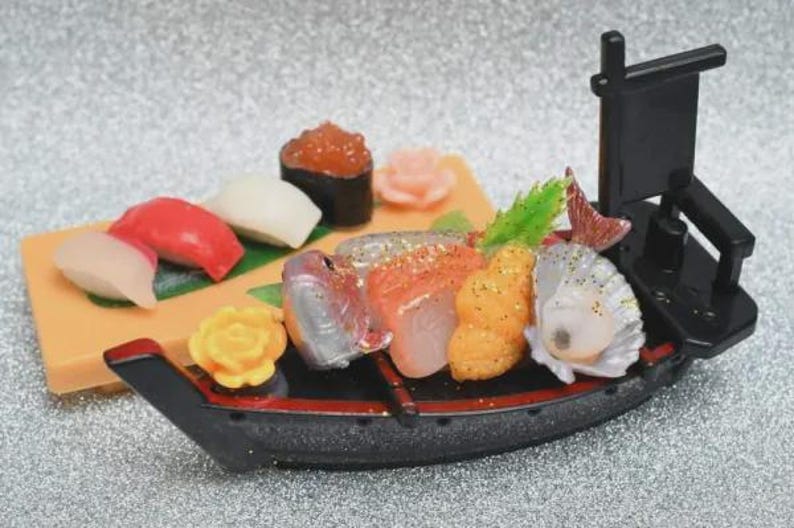 Op de afbeelding: Een miniatuur sushi boot met diverse sushi stukken, waaronder nigiri en sashimi. De boot is zwart met rode accenten, en de sushi heeft levendige kleuren zoals rood, oranje en wit. Achteraan staat een kleine zwarte vlag.