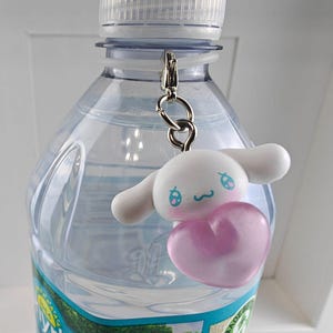 Puede incluir: Una botella de agua de pl&aacute;stico transparente con tapa blanca y un llavero plateado. El llavero presenta un personaje de dibujos animados blanco con detalles azules y un coraz&oacute;n rosa.