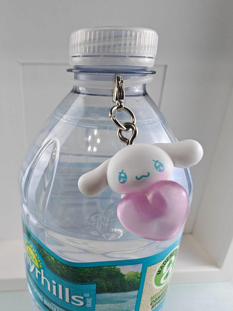 Puede incluir: Una botella de agua de pl&aacute;stico transparente con tapa blanca y un llavero plateado. El llavero presenta un personaje de dibujos animados blanco con detalles azules y un coraz&oacute;n rosa.