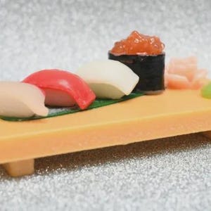 Op de afbeelding: Een miniatuur sushiplankje met diverse stukjes sushi, waaronder nigiri en een makirrol, gepresenteerd op een rechthoekig houten dienblad. De sushi bevat zalmkuit, gember en wasabi. Het dienblad is ongeveer 10 cm lang.
