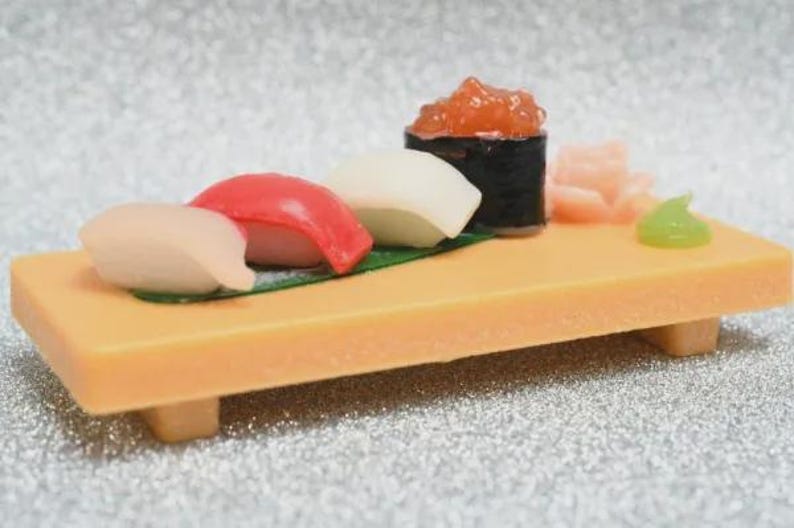 Op de afbeelding: Een miniatuur sushiplankje met diverse stukjes sushi, waaronder nigiri en een makirrol, gepresenteerd op een rechthoekig houten dienblad. De sushi bevat zalmkuit, gember en wasabi. Het dienblad is ongeveer 10 cm lang.