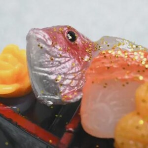 Op de afbeelding: Close-up van een decoratief sushi-arrangement met een vissenkop met rode accenten, oranje en witte visschijfjes en een gele bloem. De sushi wordt gepresenteerd op een zwarte tray met rode rand en bestrooid met gouden glitter.