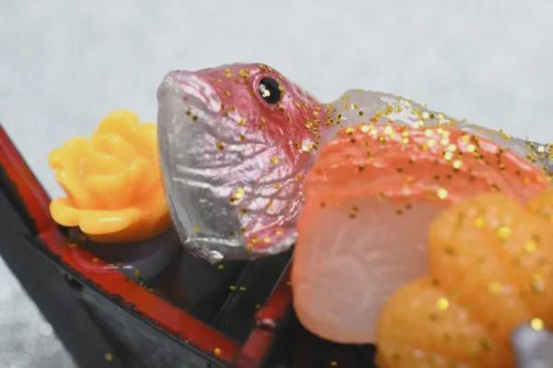 Op de afbeelding: Close-up van een decoratief sushi-arrangement met een vissenkop met rode accenten, oranje en witte visschijfjes en een gele bloem. De sushi wordt gepresenteerd op een zwarte tray met rode rand en bestrooid met gouden glitter.