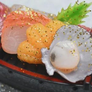 Op de afbeelding: Een miniatuur sushi boot met diverse zeevruchten. De opstelling omvat zalm, oranje kuit, een sint-jakobsschelp en een vissenkop. Alle items zijn versierd met gouden glitter. De boot is zwart met rode rand.