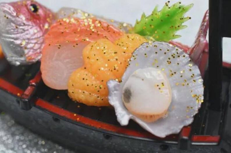 Op de afbeelding: Een miniatuur sushi boot met diverse zeevruchten. De opstelling omvat zalm, oranje kuit, een sint-jakobsschelp en een vissenkop. Alle items zijn versierd met gouden glitter. De boot is zwart met rode rand.
