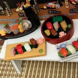 Op de afbeelding: Miniatuur sushi set met diverse nigiri en sashimi arrangementen. Inclusief een zwart bootvormig gerecht, een rode kom en een rechthoekige plaat, allemaal gevuld met kleurrijke sushi stukken. De set staat op een kleine witte tafel. Afmetingen in cm.