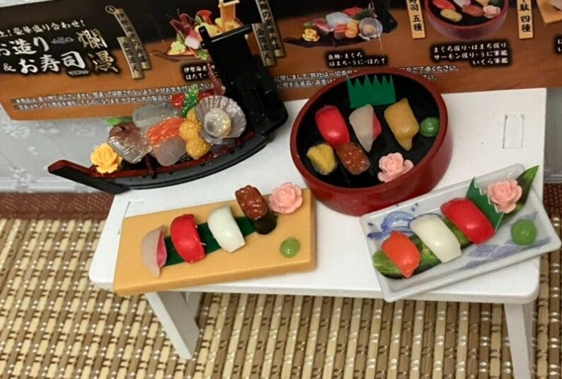 Op de afbeelding: Miniatuur sushi set met diverse nigiri en sashimi arrangementen. Inclusief een zwart bootvormig gerecht, een rode kom en een rechthoekige plaat, allemaal gevuld met kleurrijke sushi stukken. De set staat op een kleine witte tafel. Afmetingen in cm.
