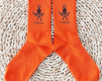 May include: A pair of bright orange socks with a black graphic of a skeleton meditating, with the word "THINKING" printed below. The socks are made of a soft, ribbed material and are displayed on a woven surface.