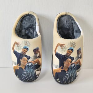 May include: A pair of beige slippers with a family portrait printed on the top. The slippers have a soft, gray interior and a black sole. The family is wearing dark blue and gray clothing.