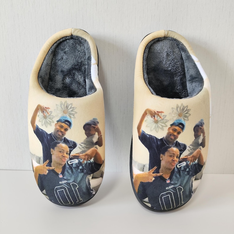 May include: A pair of beige slippers with a family portrait printed on the top. The slippers have a soft, gray interior and a black sole. The family is wearing dark blue and gray clothing.
