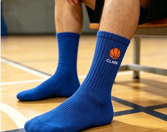 May include: A pair of royal blue crew socks featuring a ribbed texture. Each sock displays an orange basketball graphic positioned above the white text "CLARK" on the outer side of the upper ankle area. The socks are made of a solid blue fabric.