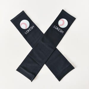 May include: A pair of black arm sleeves featuring a white baseball graphic with red stitching and the text LOKESH printed in white capital letters. The sleeves are positioned in an X shape against a plain, light-coloured background.