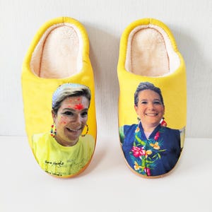 May include: A pair of yellow slippers with a soft, cream-colored interior. Each slipper features a printed image of a smiling person. One slipper has text that reads "Bone Heads" and "BOP" in a script font.