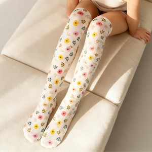 Può includere: Un paio di calze alte bianche con un motivo floreale. Le calze presentano un motivo ripetuto di fiori gialli, rosa e blu con foglie verdi. Le calze sono realizzate in un materiale morbido e leggero.