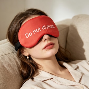 Könnte beinhalten: Eine leuchtend rote Schlafmaske mit dem weißen Schriftzug "Do not disturb" auf der Vorderseite. Die Maske verfügt über ein schwarzes elastisches Band, das für einen sicheren Sitz am Kopf sorgt und das Licht für Ruhe abhält.